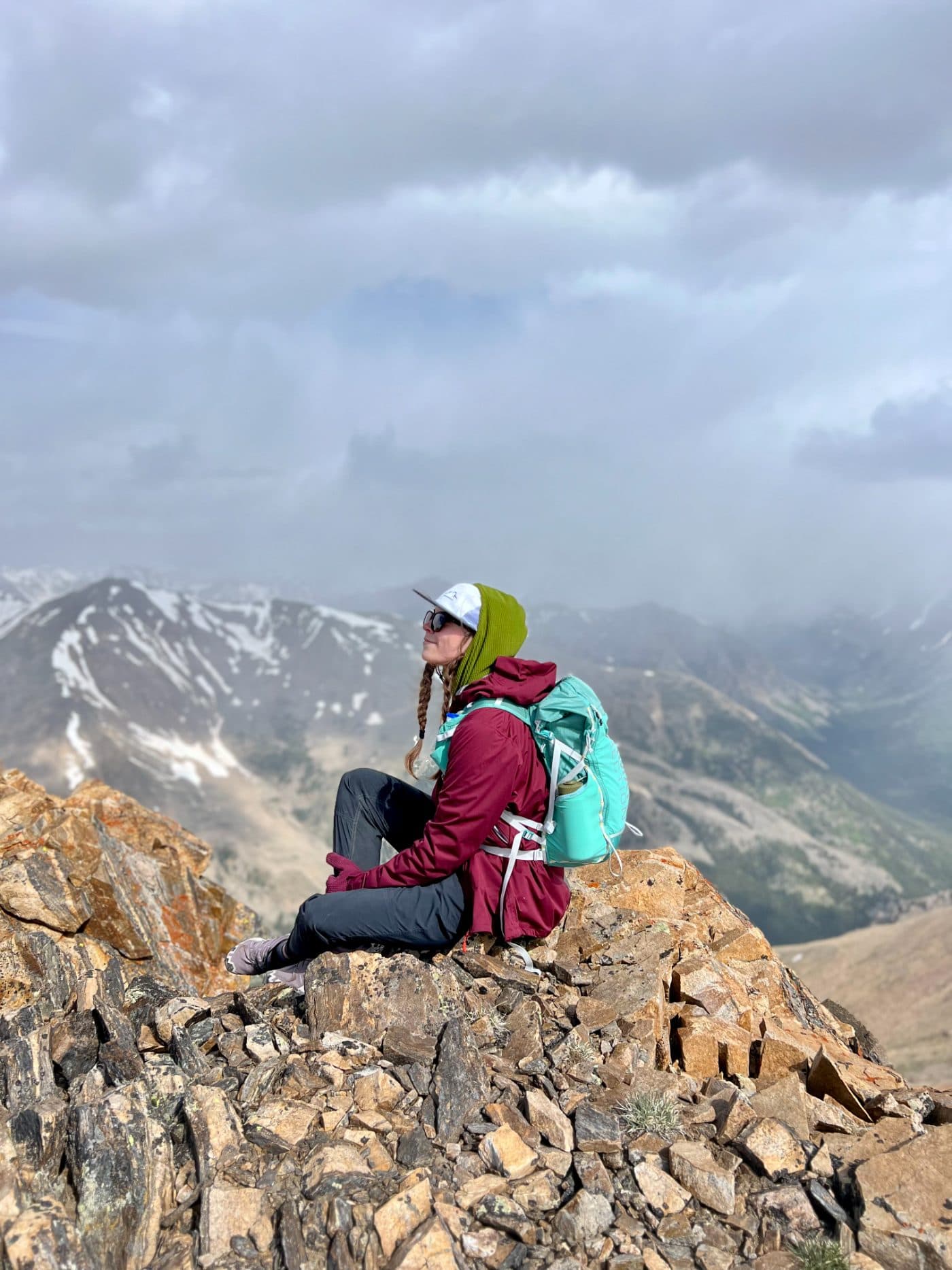 A hiker with a turquoise backpack sits on a rocky summit above snow-streaked mountains.