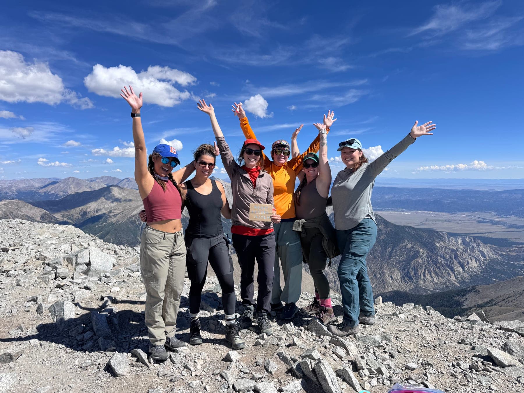 Six hikers celebrate together on a rocky summit with mountains behind them.