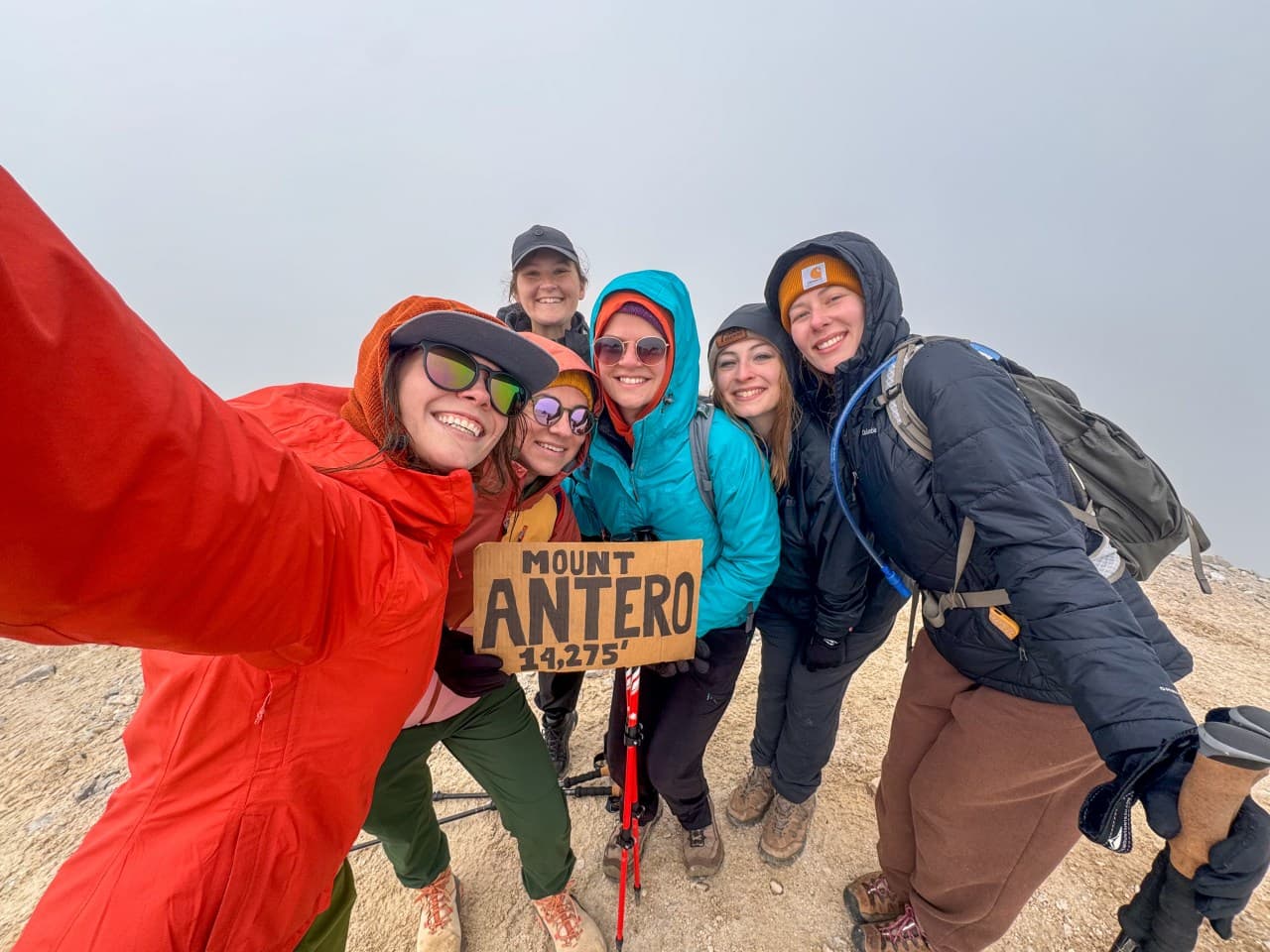 Six hikers in colorful jackets pose together with a Mount Antero summit sign.