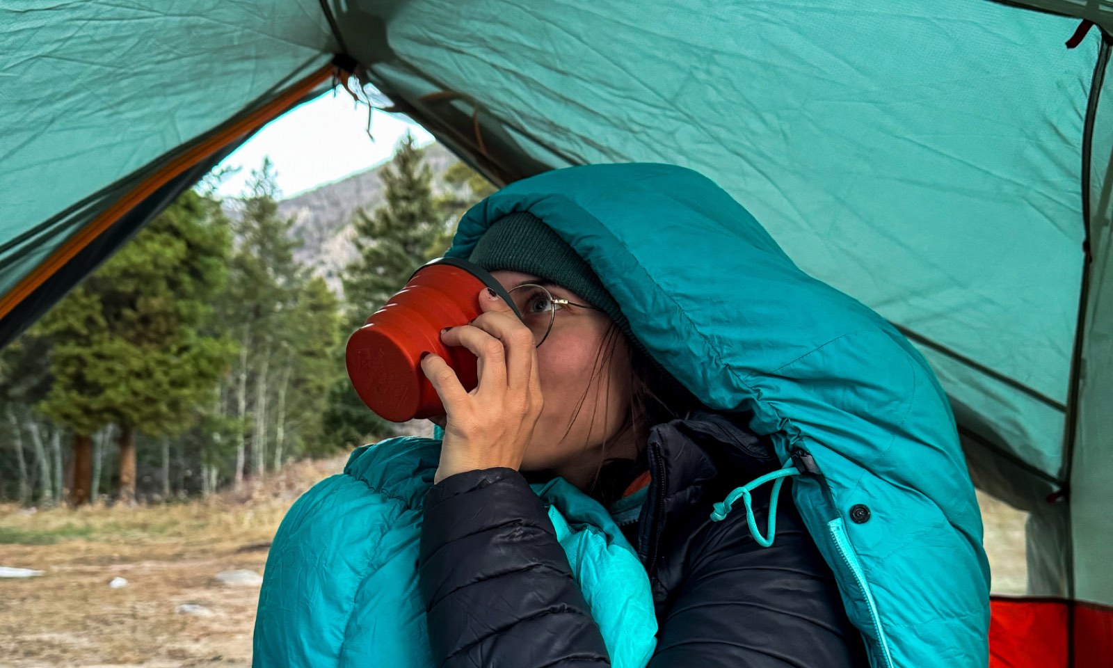 A hiker wrapped in a sleeping bag inside a green tent, sipping from a red collapsible cup, with pines and a mountainside through the open door.