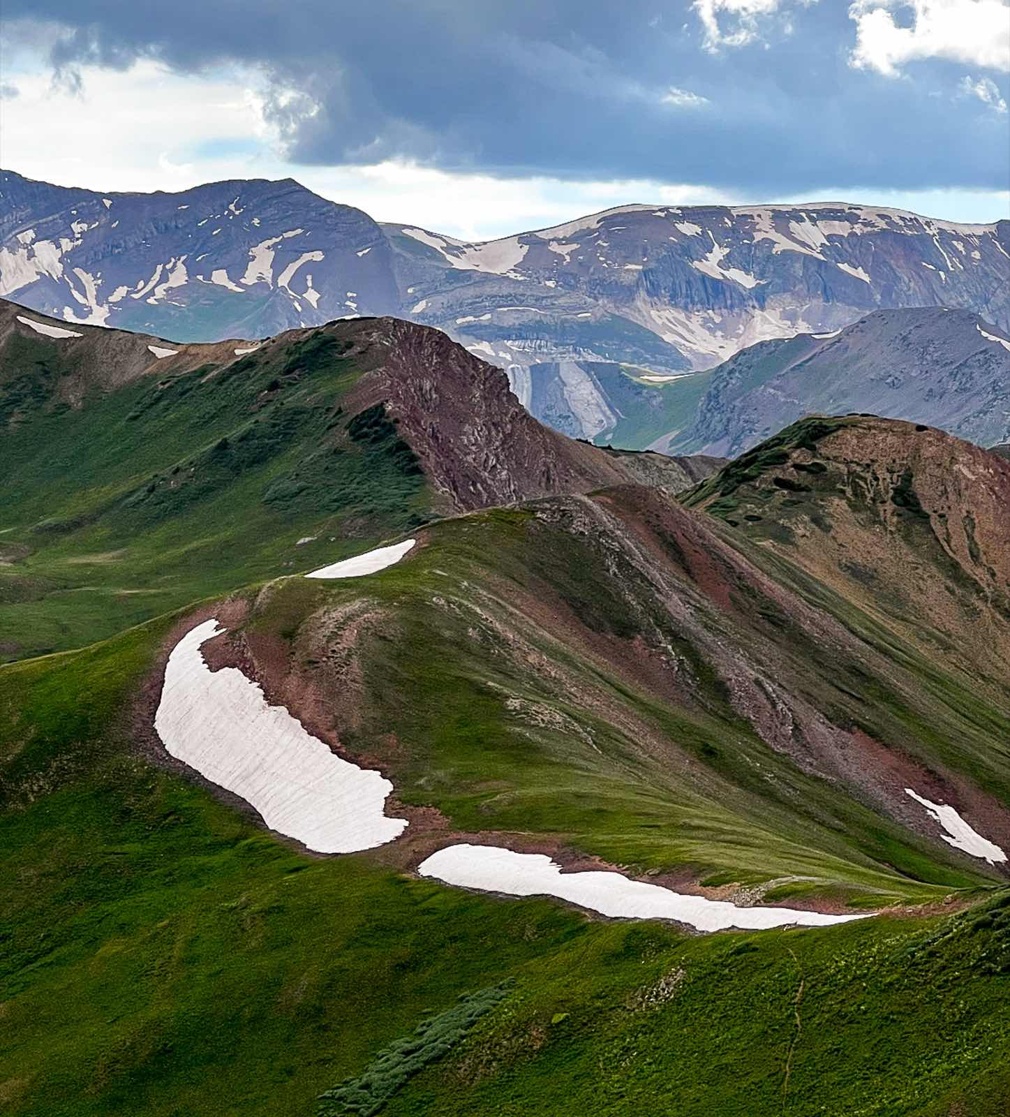 A high alpine ridgeline dotted with lingering snowfields, with layers of Colorado mountains fading into the distance under a cloudy sky.