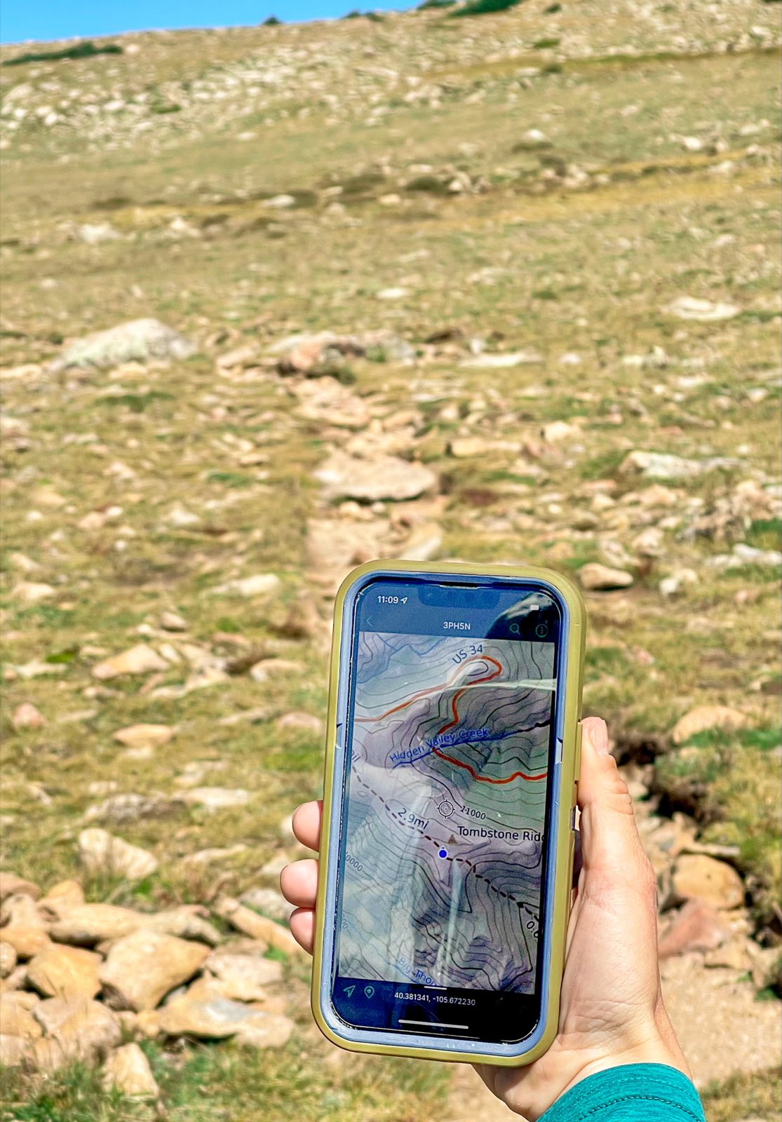 A hiker holding up a phone displaying a topographic navigation app on a rocky, high-alpine trail.
