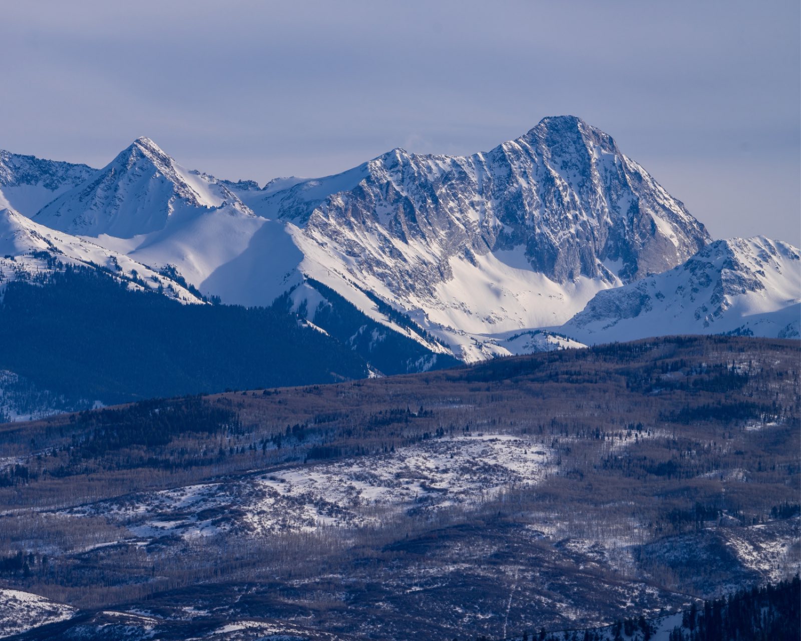 Snow-covered Colorado peaks rising above a valley dusted with fresh snow under a hazy winter sky.