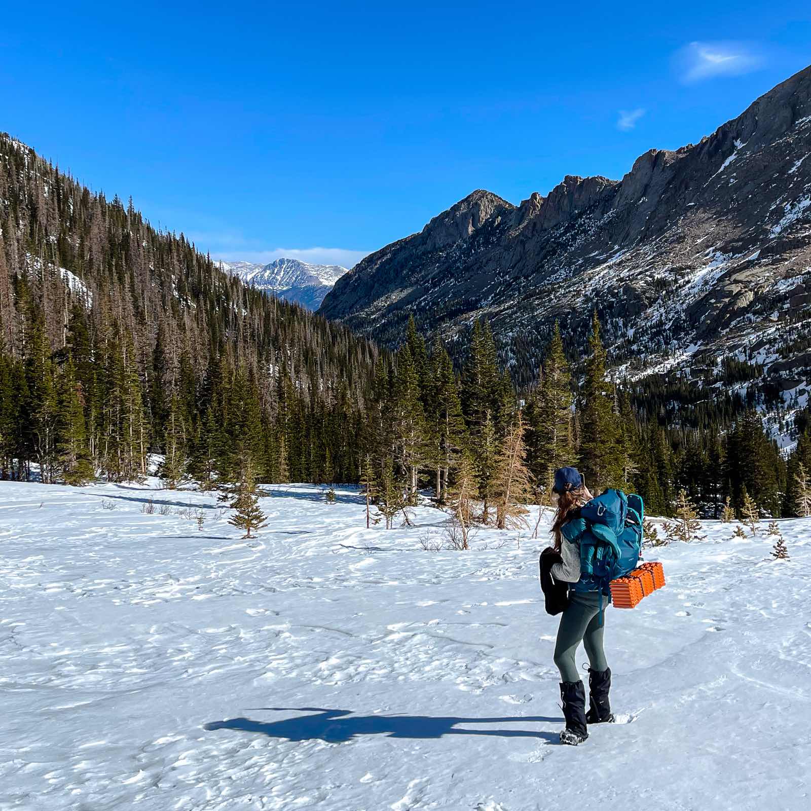 A backpacker in winter gear looking out across a snow-covered valley toward a rugged, snow-dusted mountain range.