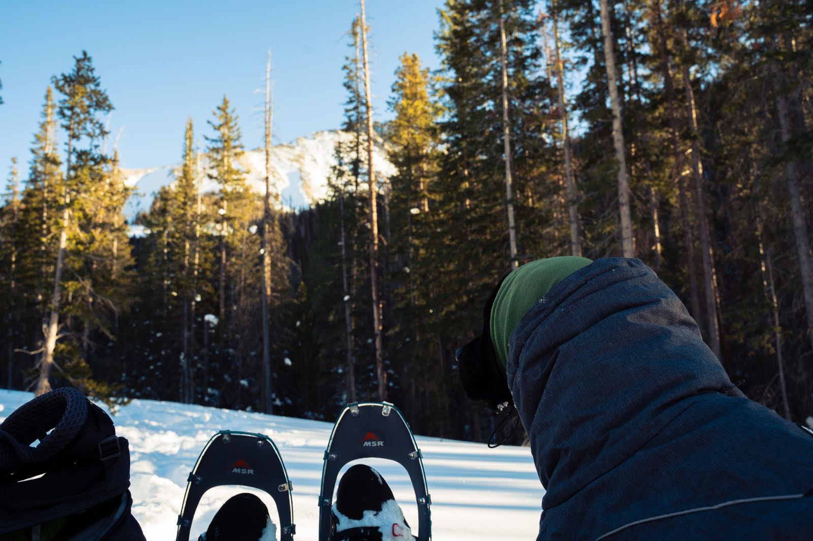 A snowshoer's MSR snowshoes and hooded jacket in the foreground, with sunlit pines and a snowy peak behind.