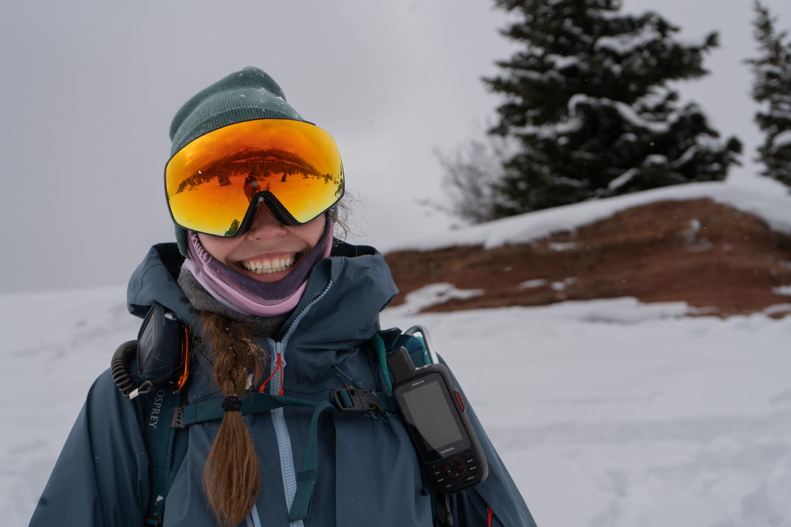 A smiling winter hiker in a hooded shell jacket, ski goggles, and a pack-mounted radio and GPS unit, standing on a snowy overlook.