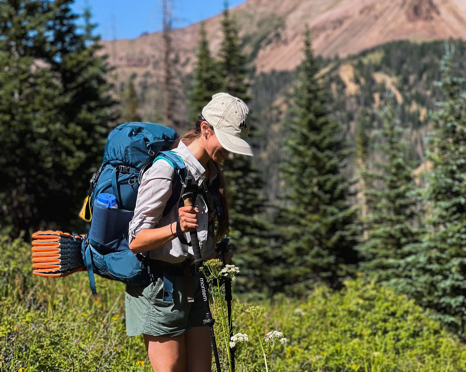 A backpacker with a loaded pack and trekking poles pausing in a wildflower meadow, mountains rising behind her.