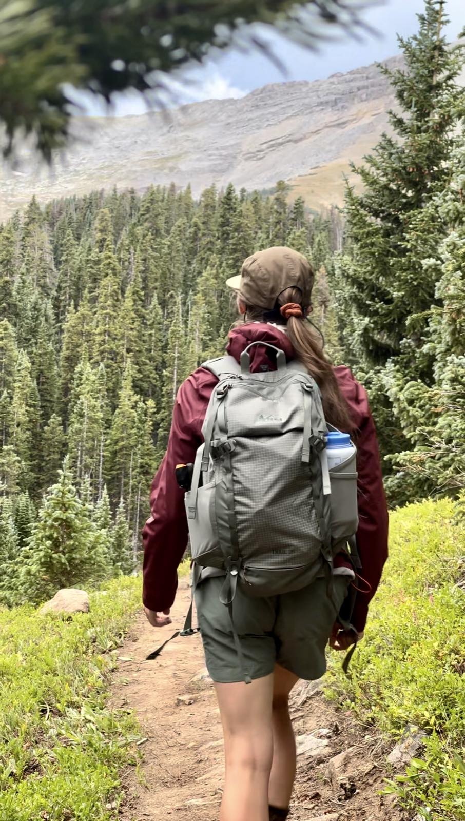 A hiker in a maroon rain jacket and gray daypack walking a forest trail toward a rocky, snow-streaked ridge.