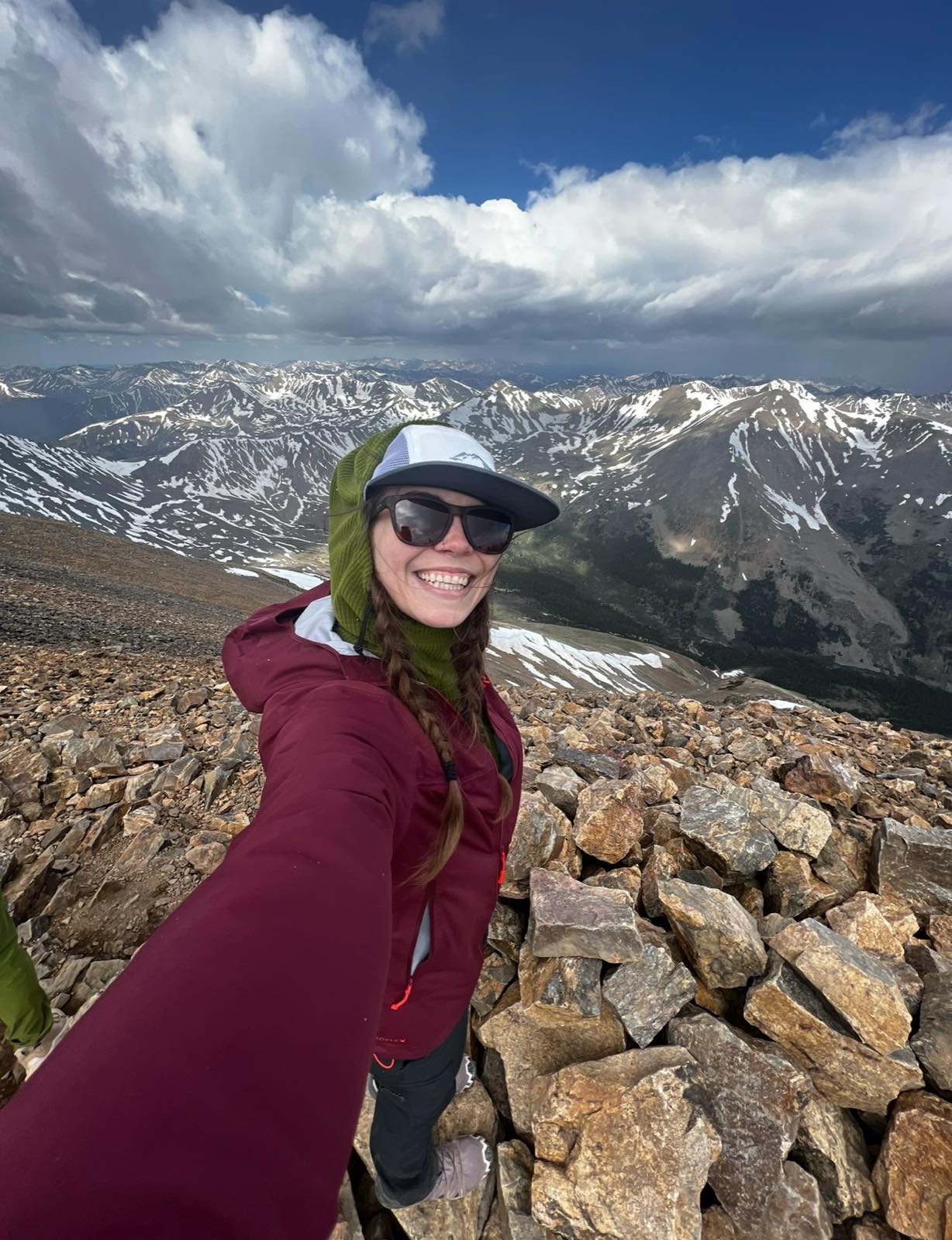 A smiling hiker in a rain jacket and sun hat taking a selfie on a rocky summit, with a sweeping view of snow-streaked mountains behind her.