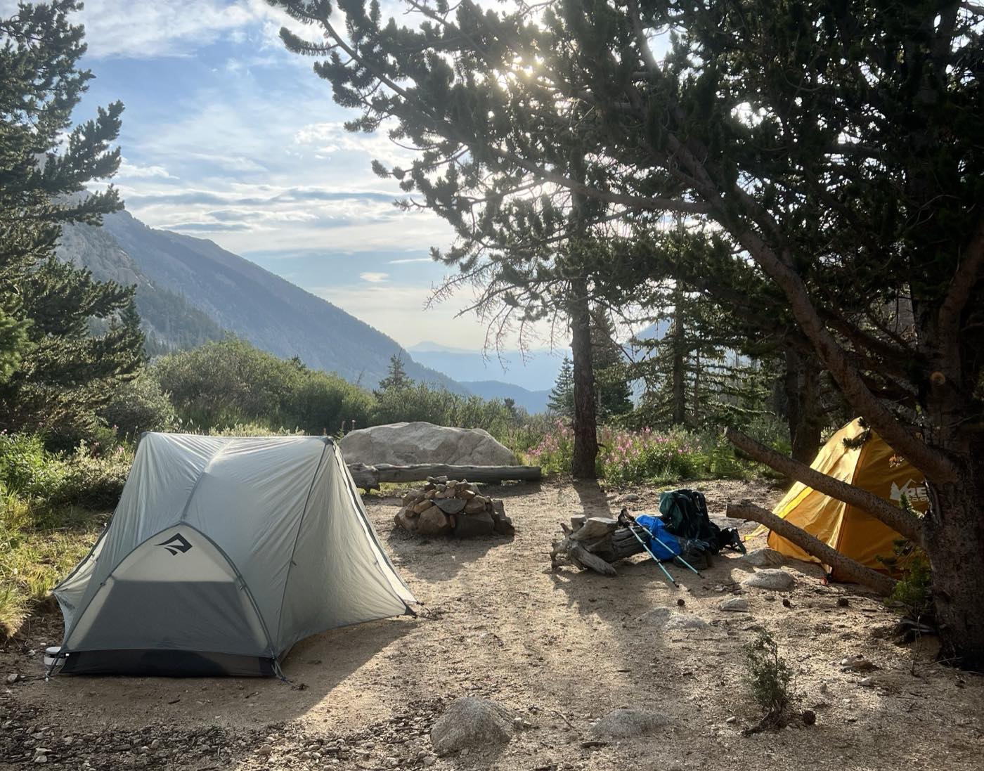 Two backpacking tents pitched in a mountain clearing at sunrise, packs and trekking poles resting against a fallen log beside a stone fire ring.