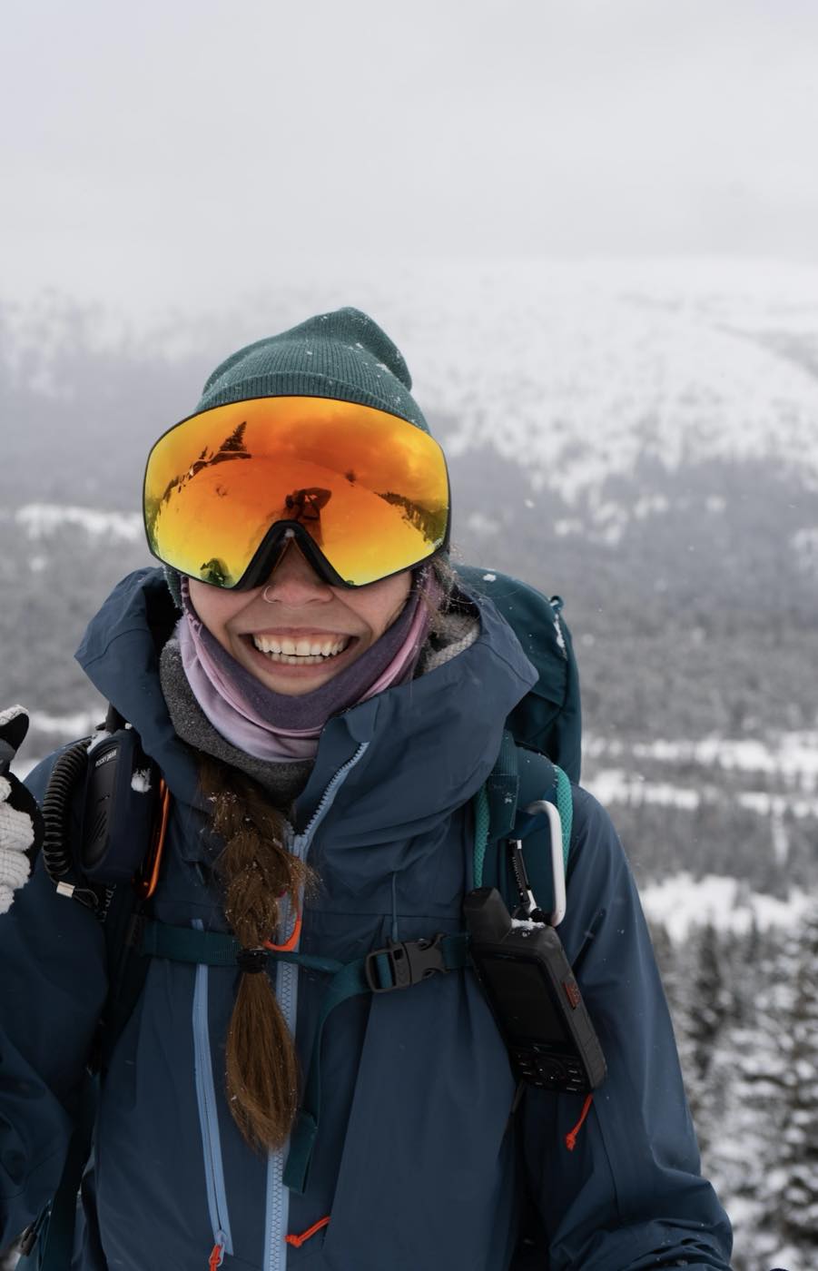A hiker in a red down jacket walks a snowy alpine trail below rugged, snow-covered peaks.