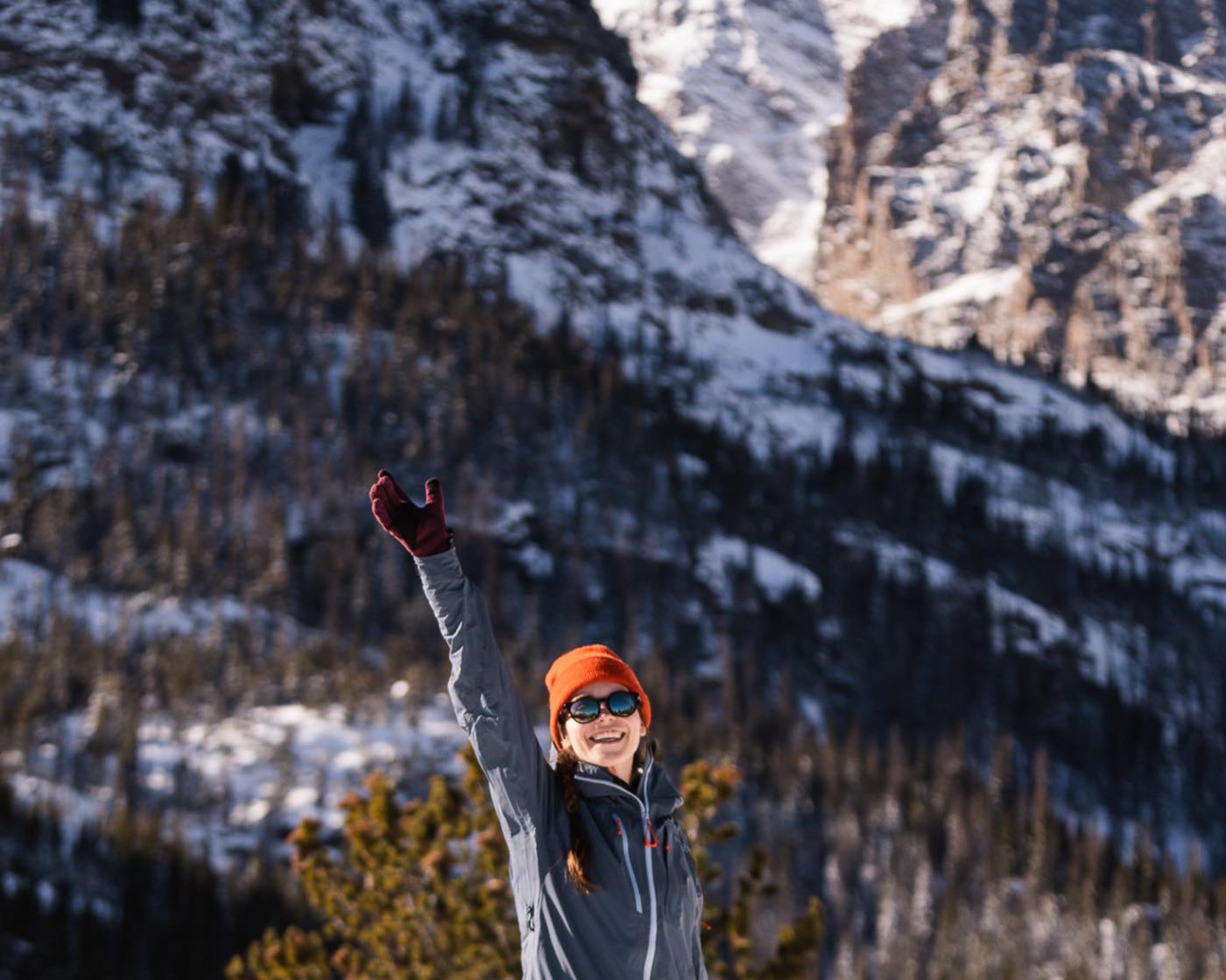 A hiker in an orange beanie raises an arm in celebration against a backdrop of a snowy ridge and forest.