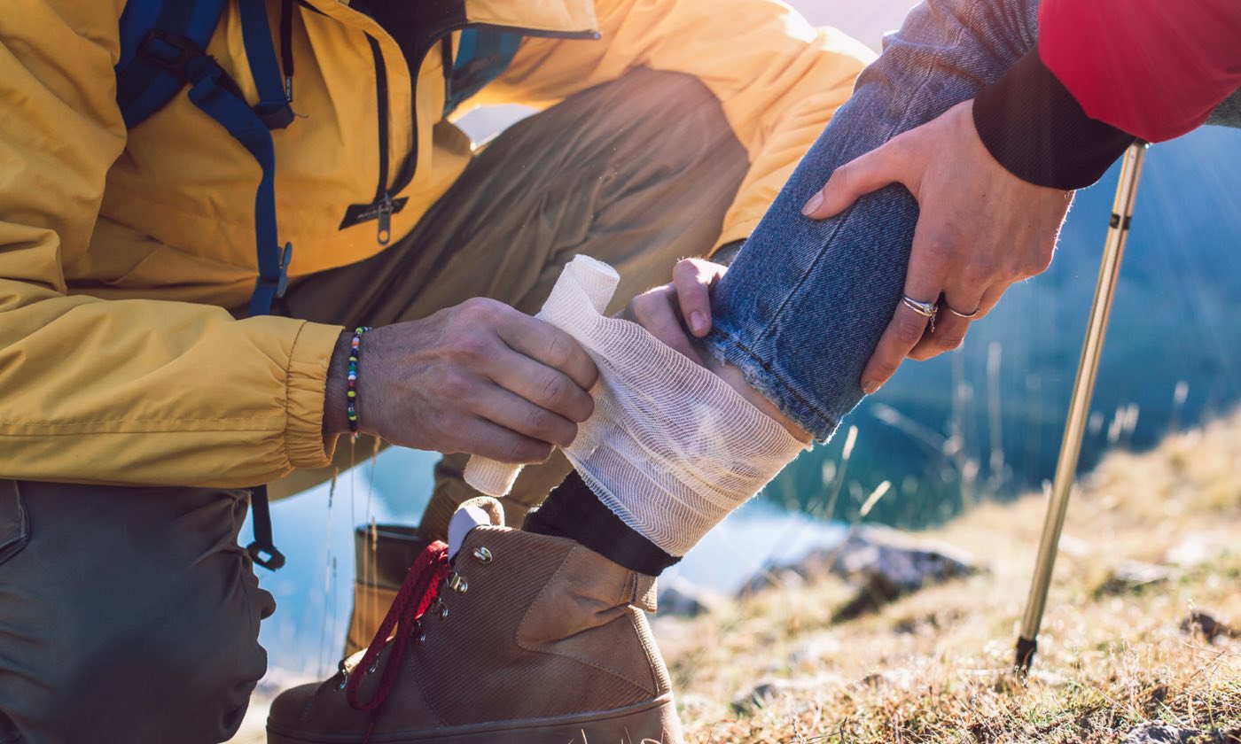A hiker wraps a gauze bandage around another hiker's lower leg on a mountainside.