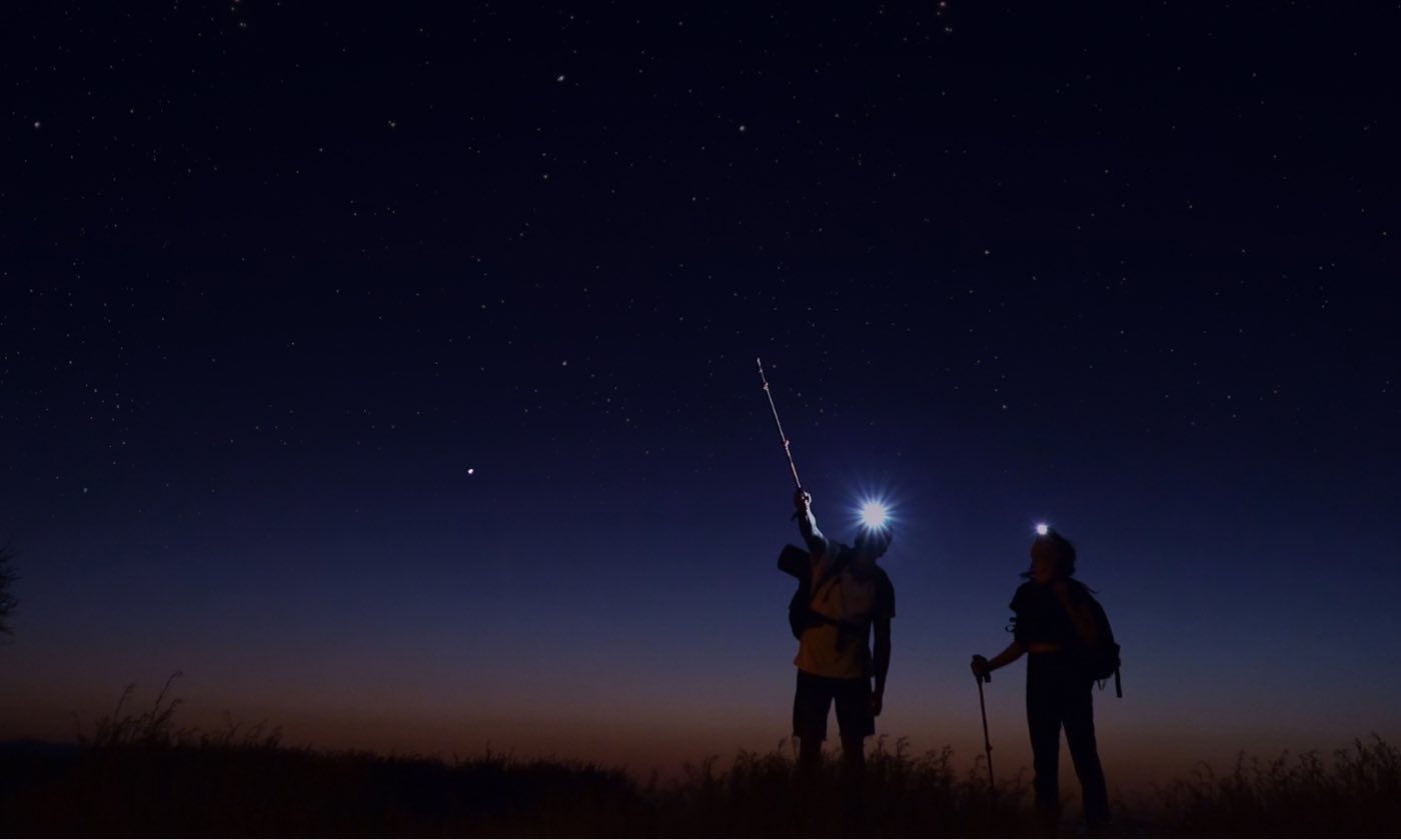 Two hikers with headlamps stand under a starry night sky, one pointing a trekking pole toward the stars.