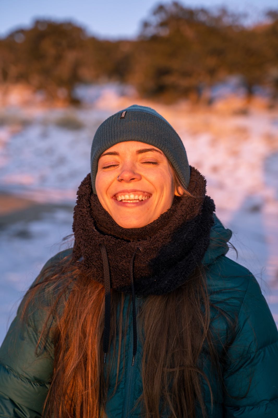 A close-up portrait of a hiker bundled in a beanie and fleece-lined hood, smiling with eyes closed in golden winter light.