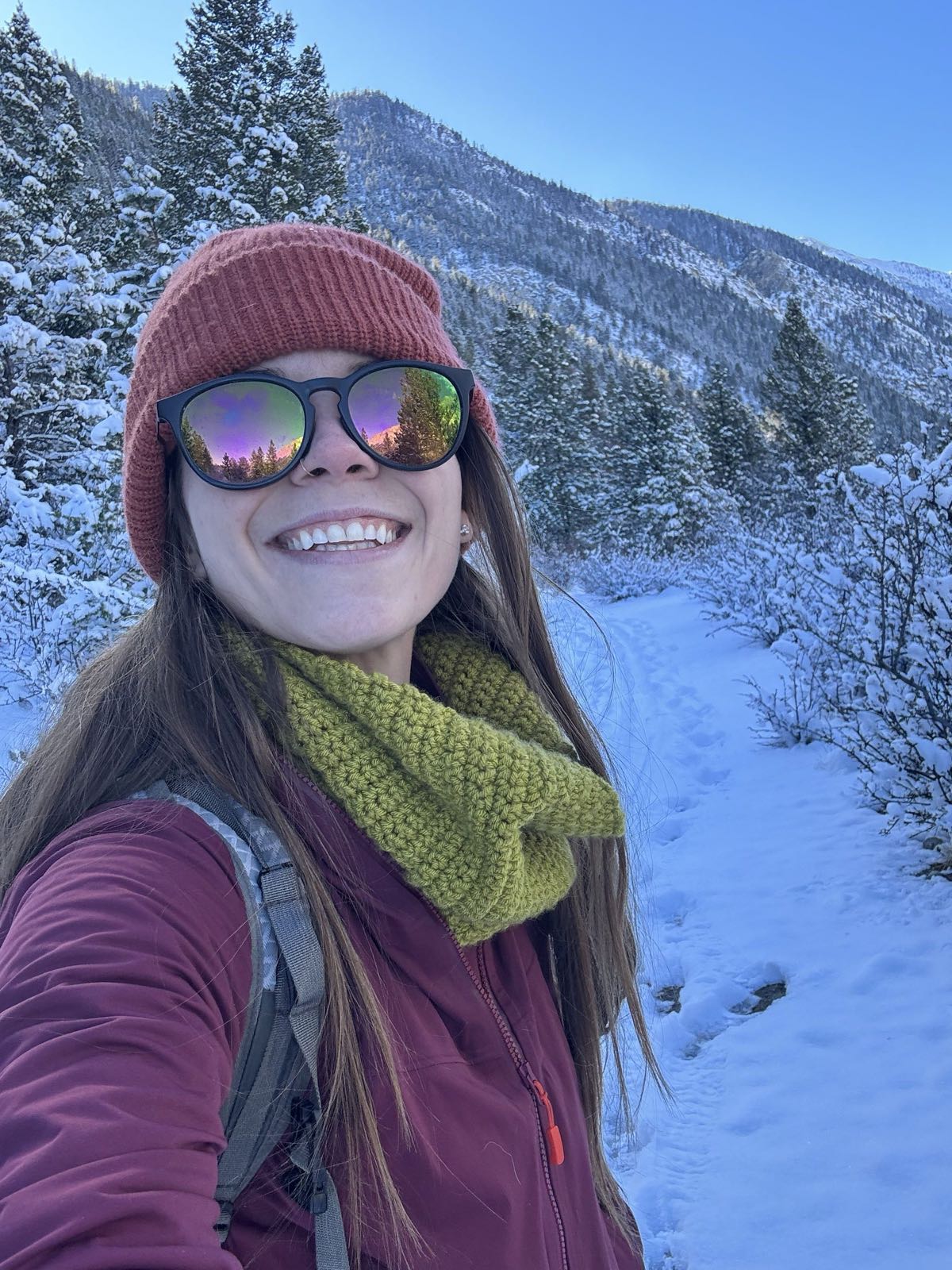 A hiker in a beanie and mirrored sunglasses smiles on a sunny snow-covered trail with mountains behind her.
