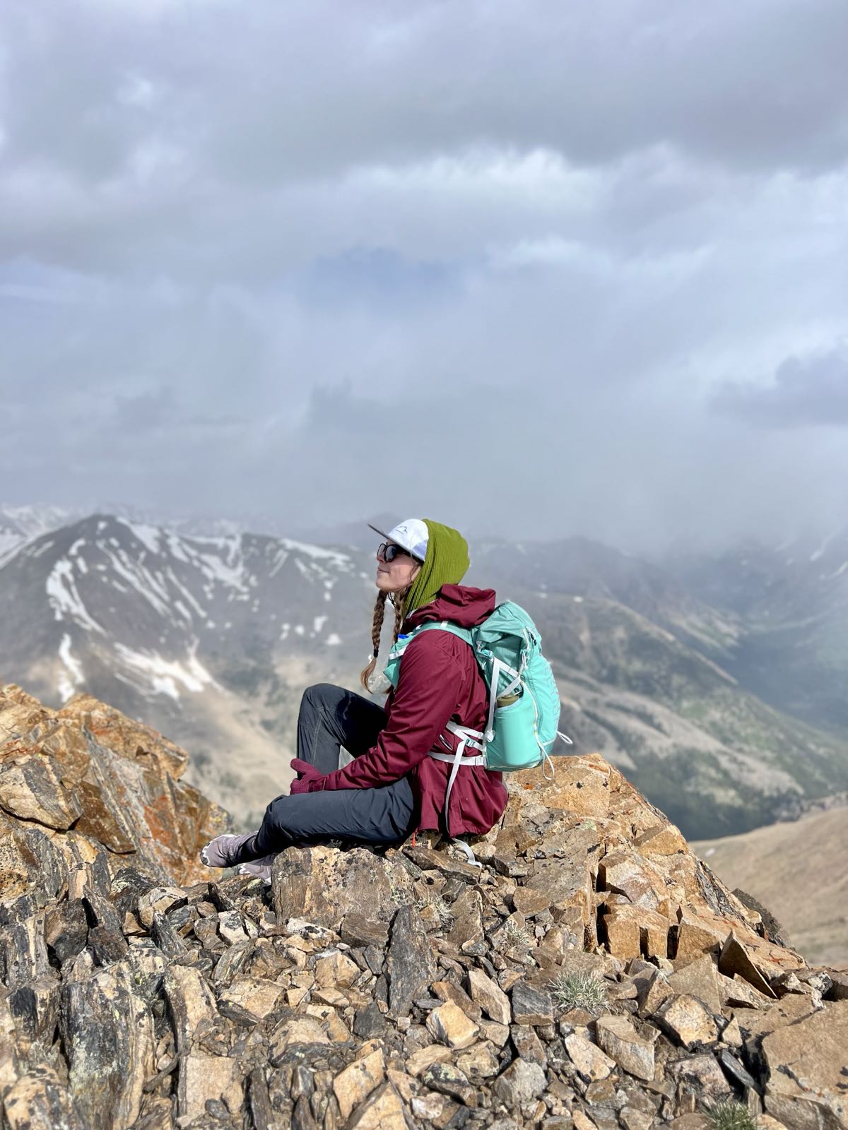 A hiker with a teal backpack sits on a rocky summit above cloud-covered peaks, looking out at the view.