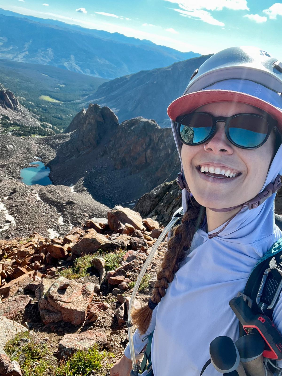 A hiker wearing sunglasses and a climbing helmet smiles on a ridge above a turquoise alpine lake and rocky basin.