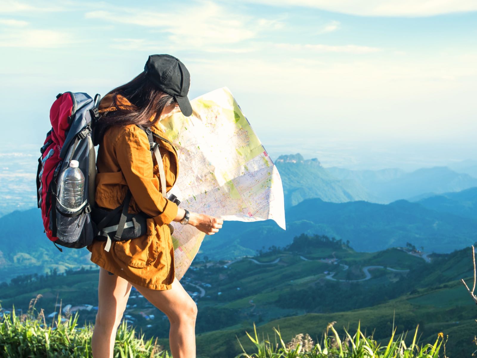 A hiker with a red backpack studies a paper map on a hilltop overlooking a green valley.