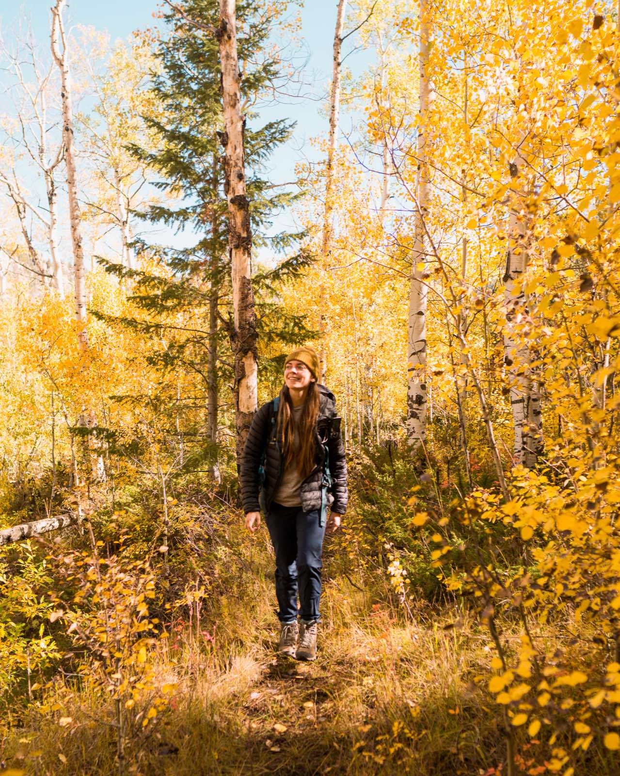 A hiker in a beanie smiles while walking a trail through golden aspen trees in fall.