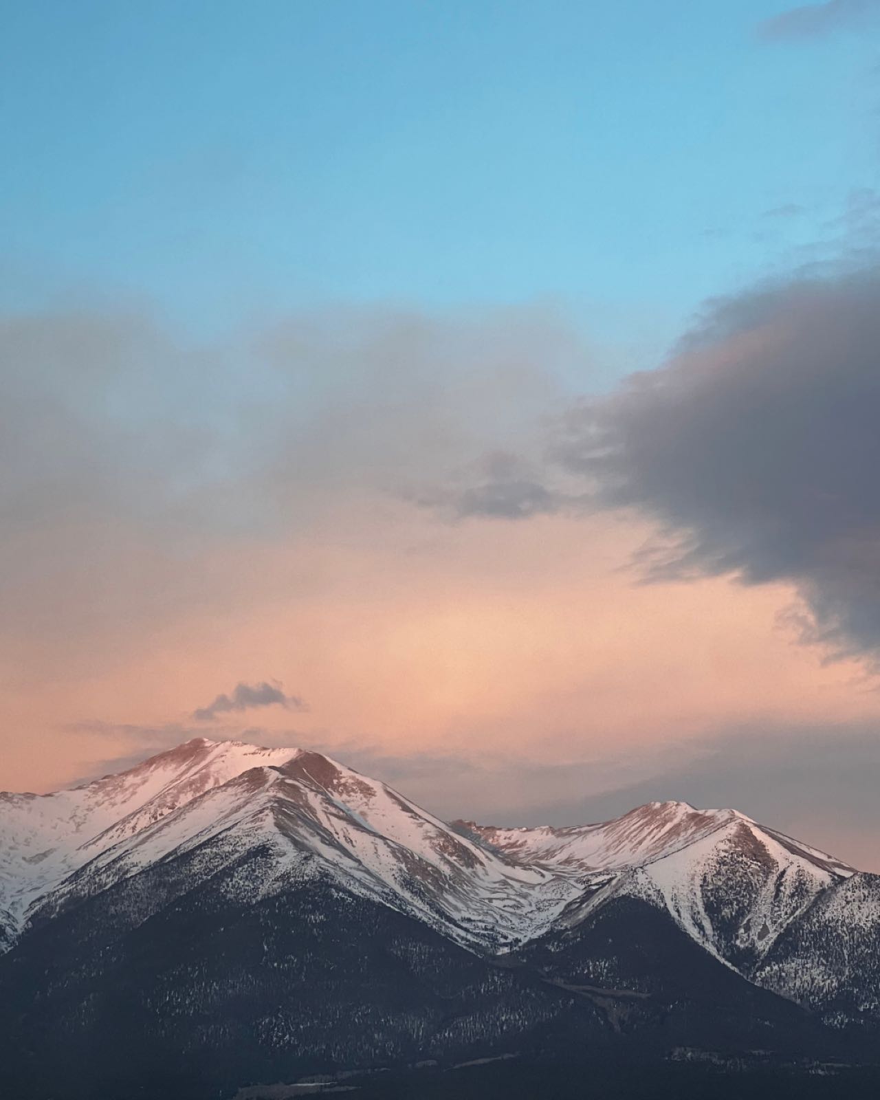A snow-capped mountain ridge glows pink and orange at sunset under a partly cloudy sky.