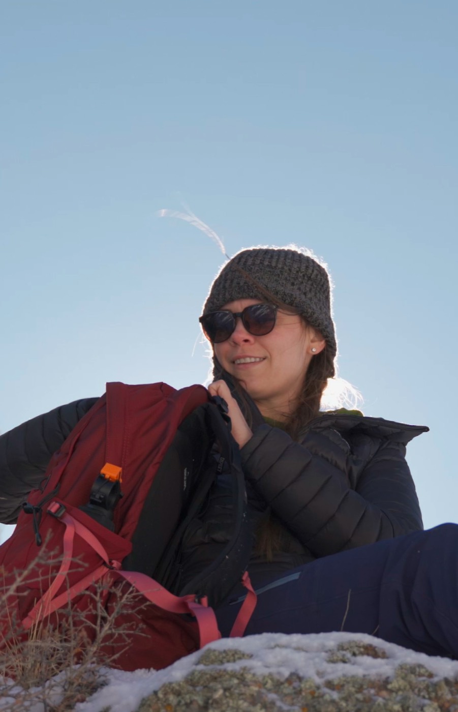 A hiker in a beanie and puffy jacket sitting on a rocky ridge with a small red daypack.