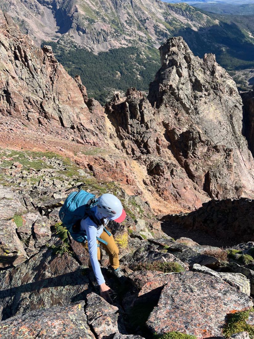 A hiker in a helmet with a teal daypack scrambling along an exposed rocky ridge.
