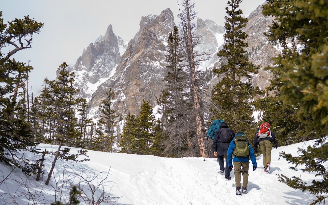 A group of hikers with packs walking a snowy trail toward jagged, snow-covered peaks.