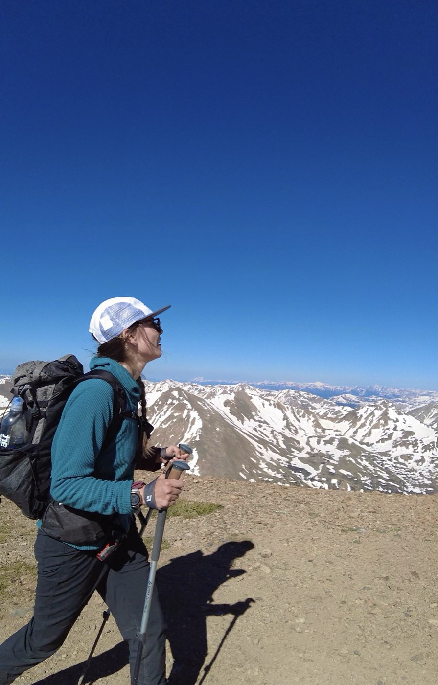 A hiker in a light trucker cap and sunglasses looking out over snow-streaked mountains with trekking poles in hand.