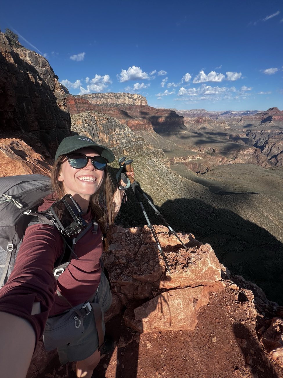 A hiker in a cap and sunglasses taking a selfie above a canyon with trekking poles and a backpack.
