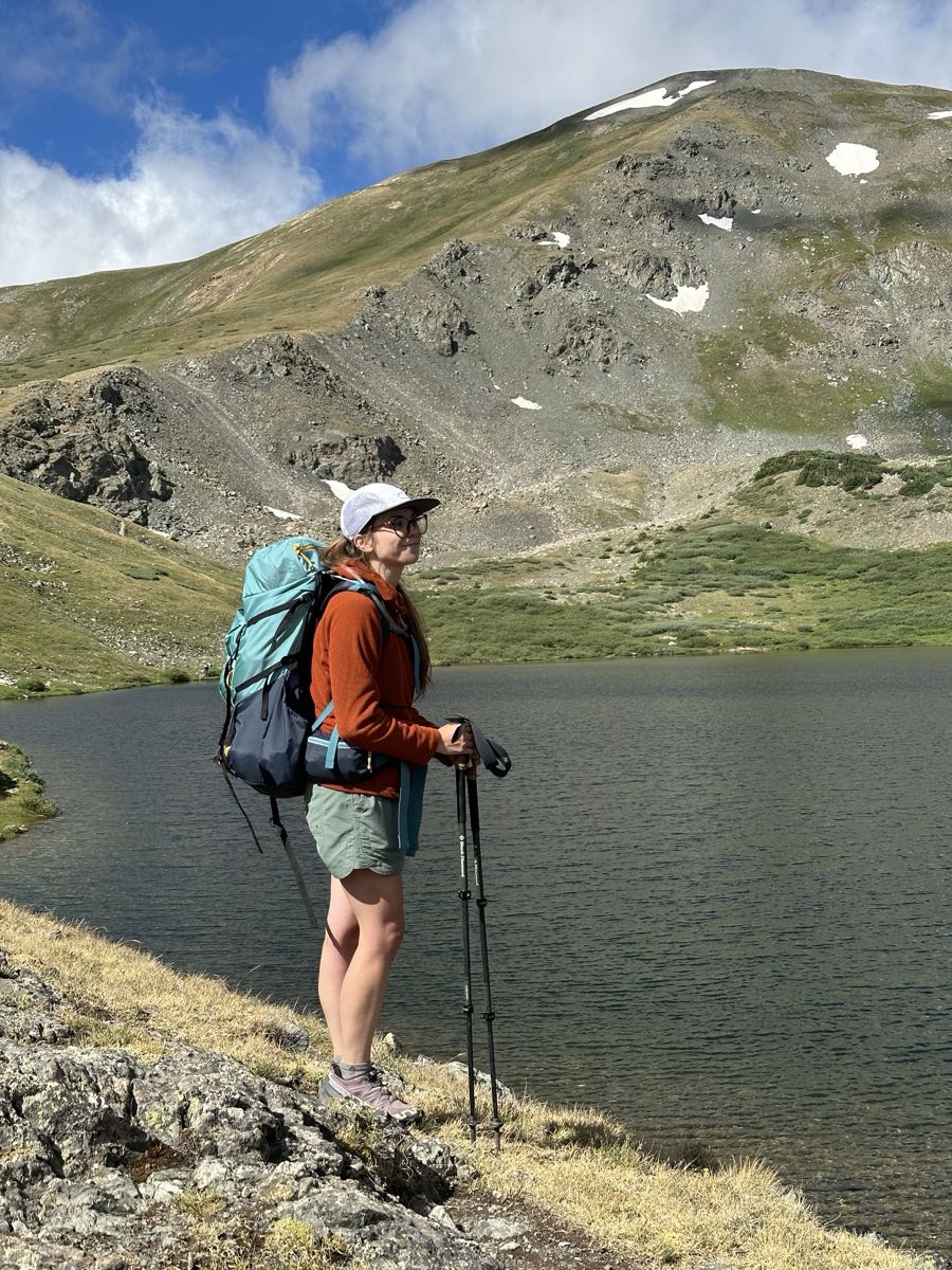 A hiker with a backpack and trekking poles standing beside an alpine lake below a snow-streaked mountain.