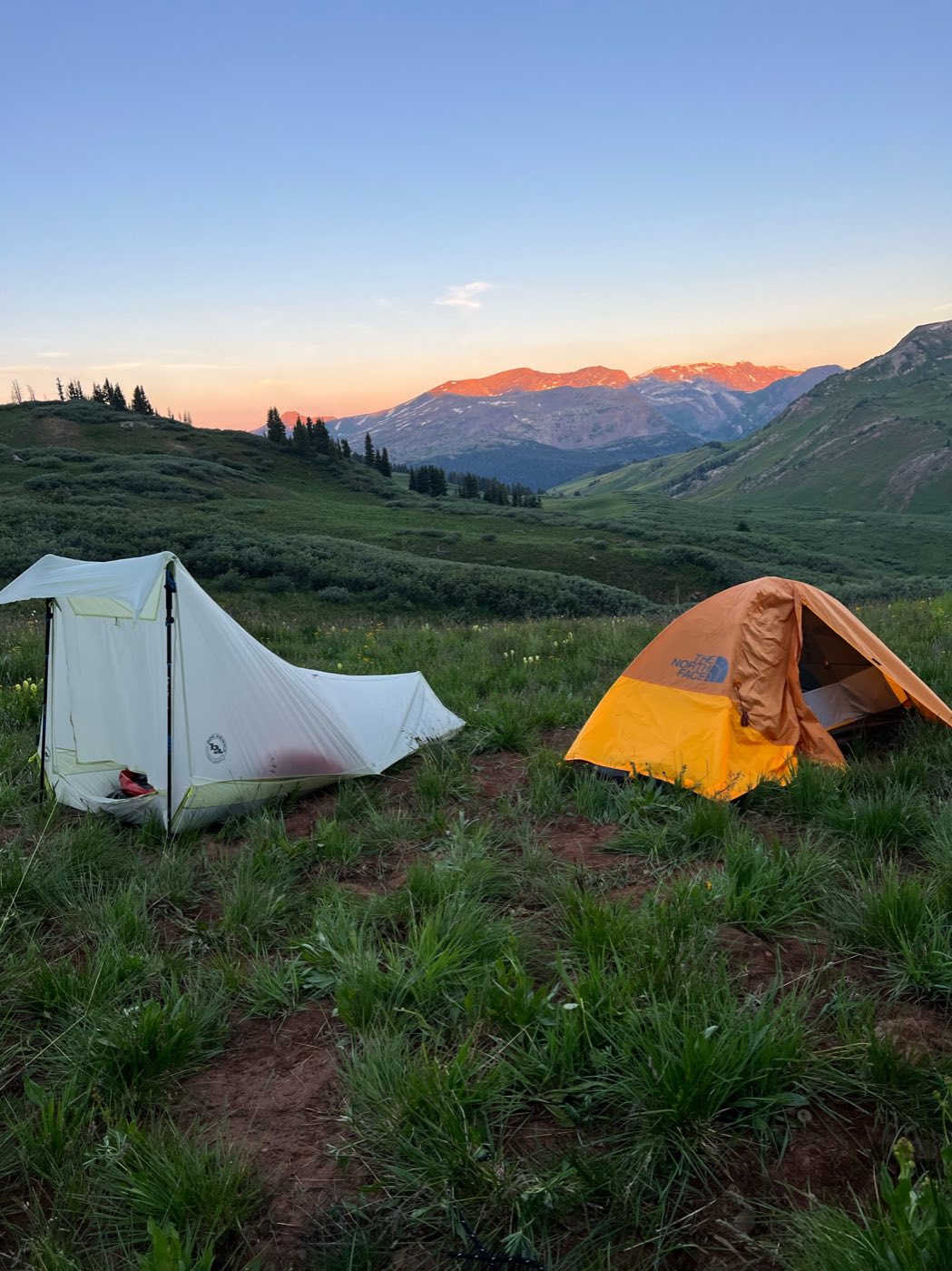 Two backpacking tents pitched in an alpine meadow at sunset, mountains glowing pink in the distance.