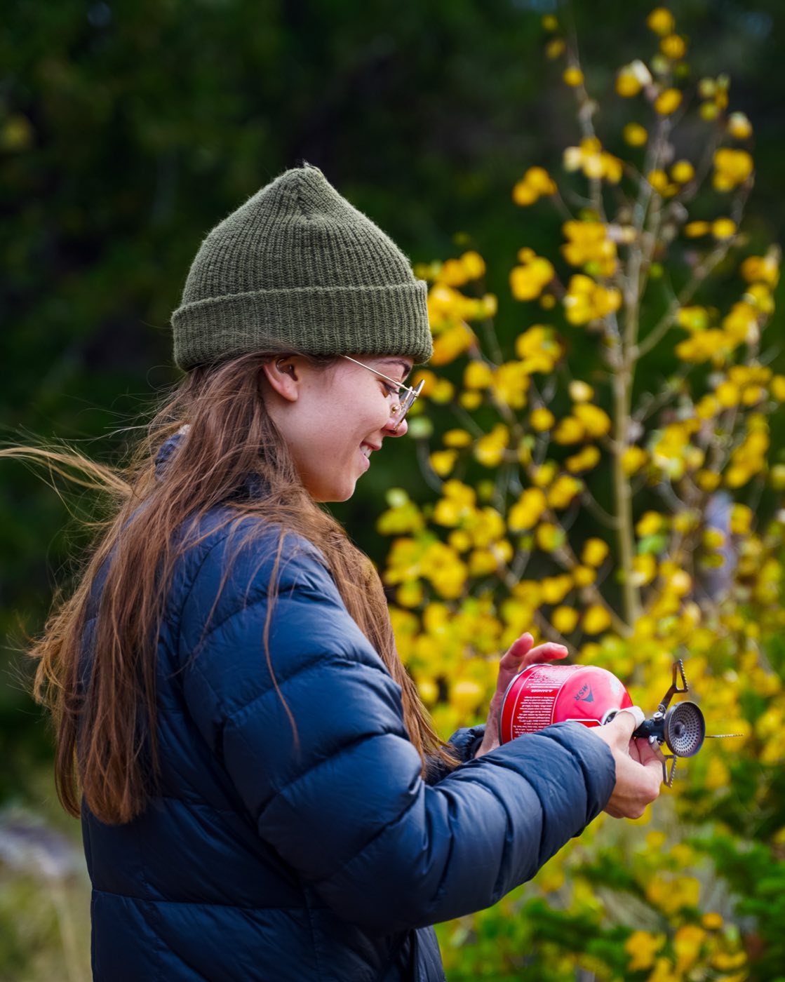 A hiker in a beanie and glasses lighting an MSR backpacking stove outdoors near autumn-yellow shrubs.