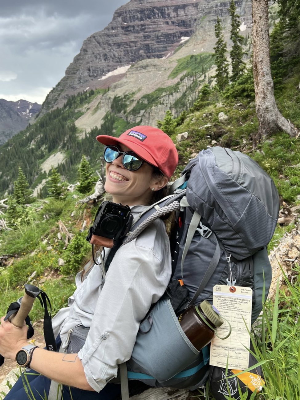 A smiling hiker in a red cap and sunglasses wearing a loaded backpacking pack with a wilderness-permit tag attached, mountains behind her.