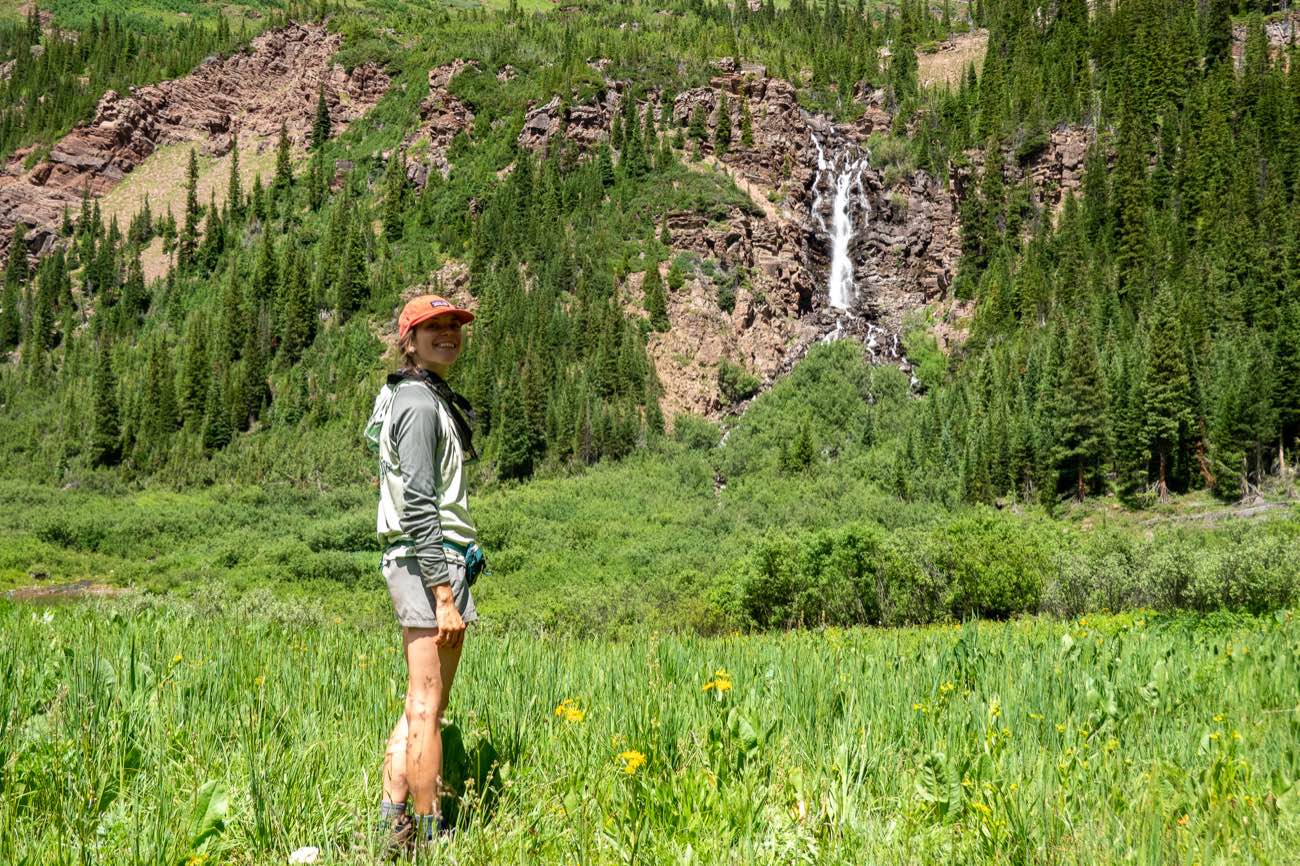 A hiker in a cap smiling in a green meadow with a waterfall cascading down a rocky hillside behind her.