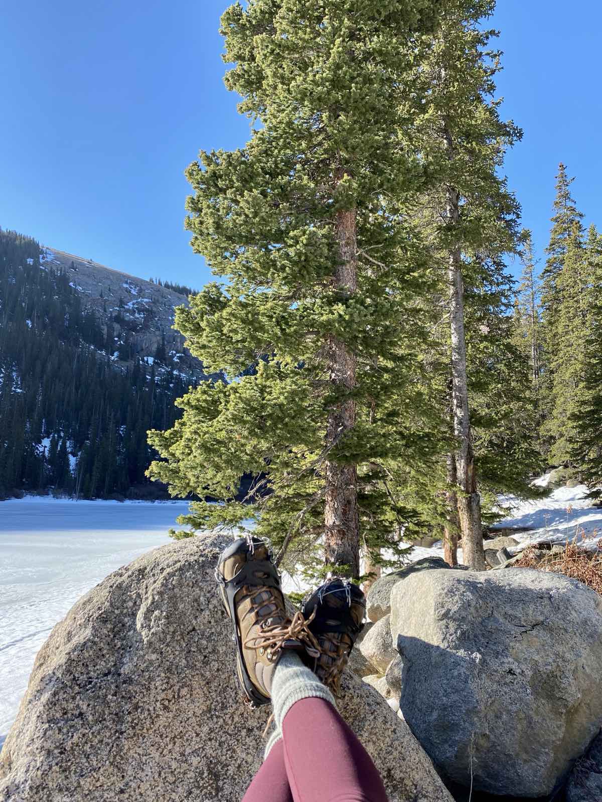Hiking boots fitted with microspikes rest on a boulder beside a frozen alpine lake.