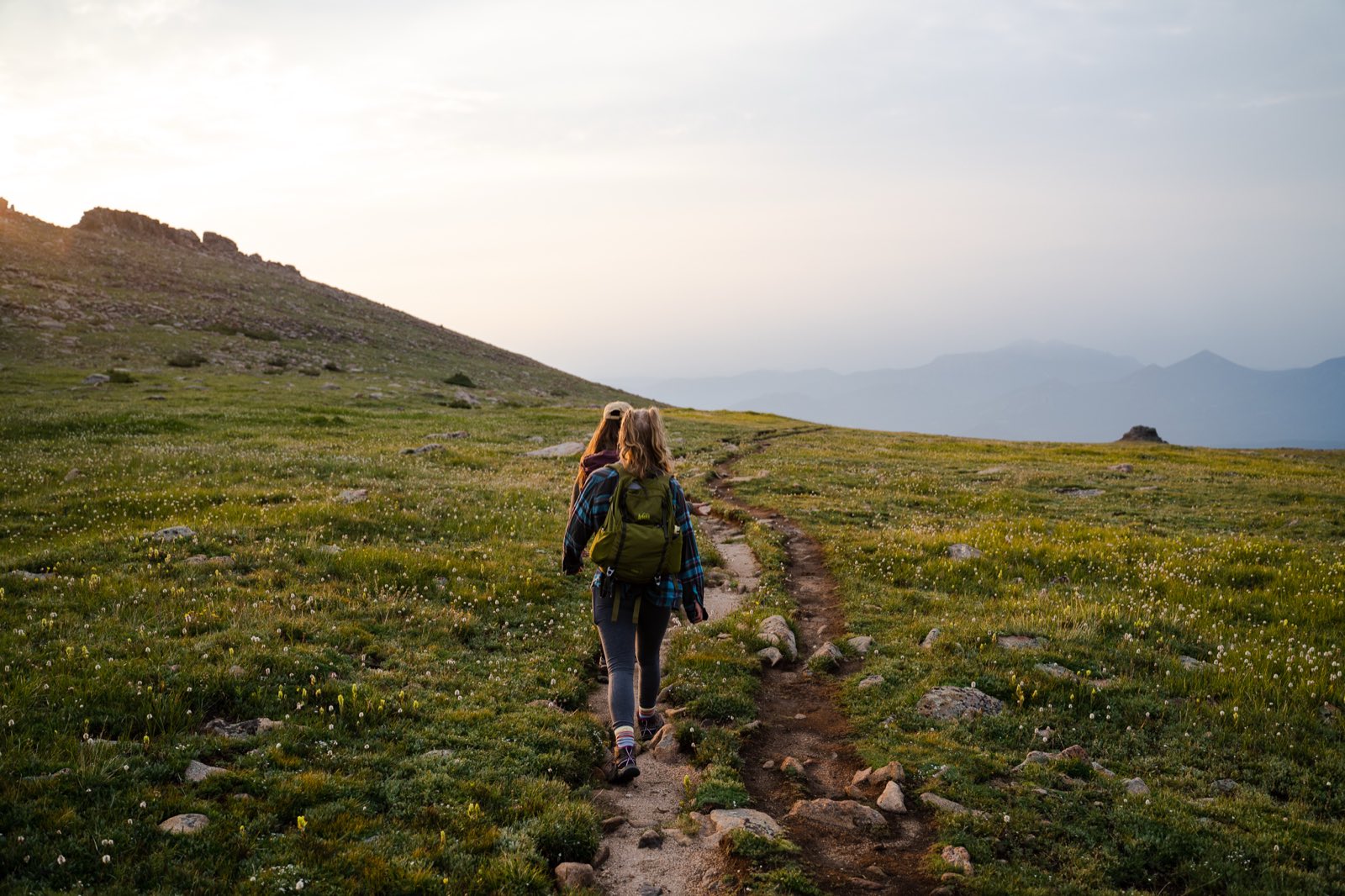 Two hikers walk a rocky tundra trail toward distant mountains at sunrise.