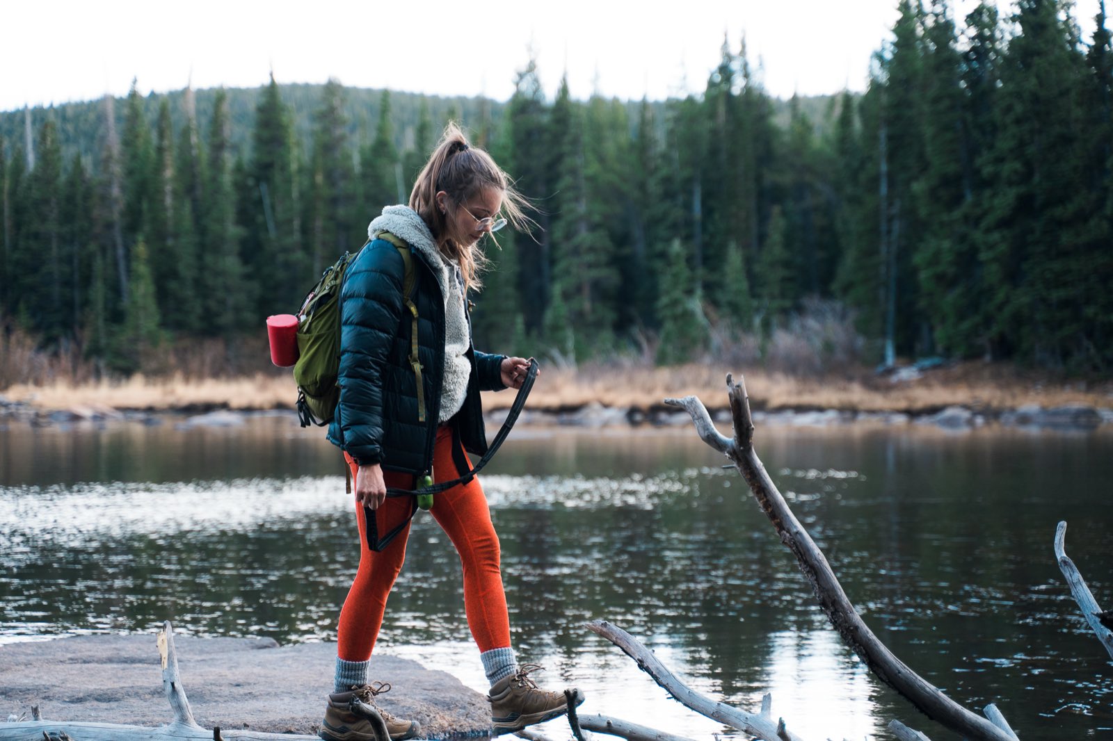 A hiker in a puffy jacket crosses a fallen log along a forested lakeshore.