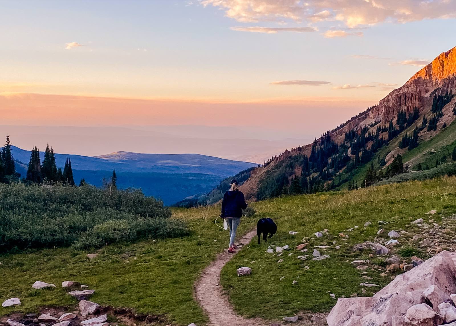 A hiker and a black dog walk a mountain trail together at golden hour.