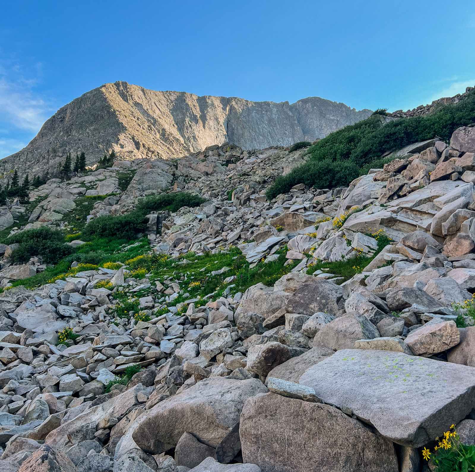 A hiker in a red cap smiles beside a backpacking partner on a wildflower trail with the Maroon Bells behind them.