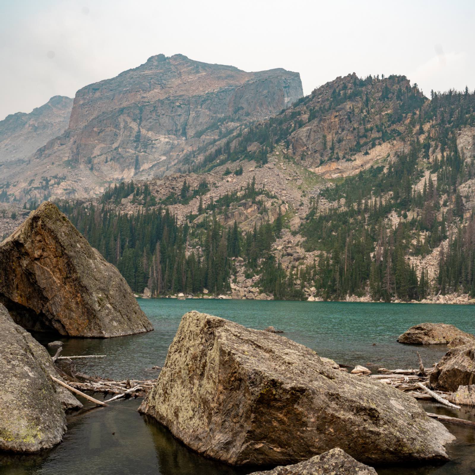 A teal alpine lake ringed by boulders and driftwood, with a hazy mountain wall rising beyond the treeline.