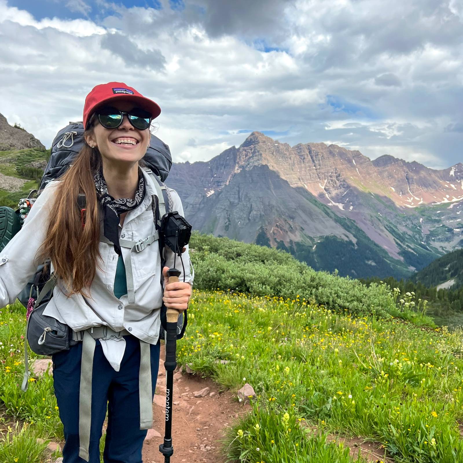 A backpacker in a red cap and sunglasses smiles on a wildflower trail with jagged red peaks in the distance.