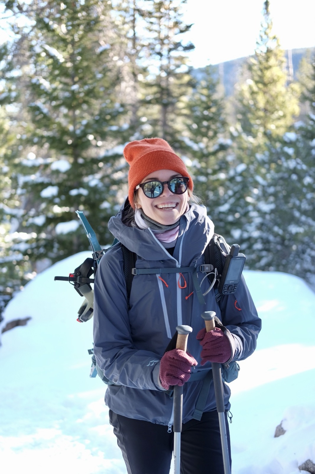 A hiker in an orange beanie and gray puffy jacket smiles on a snowy trail, trekking poles in hand.