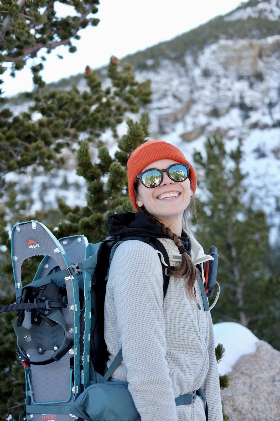 A hiker in an orange beanie and sunglasses smiles with snowshoes strapped to her pack, snowy peaks behind her.