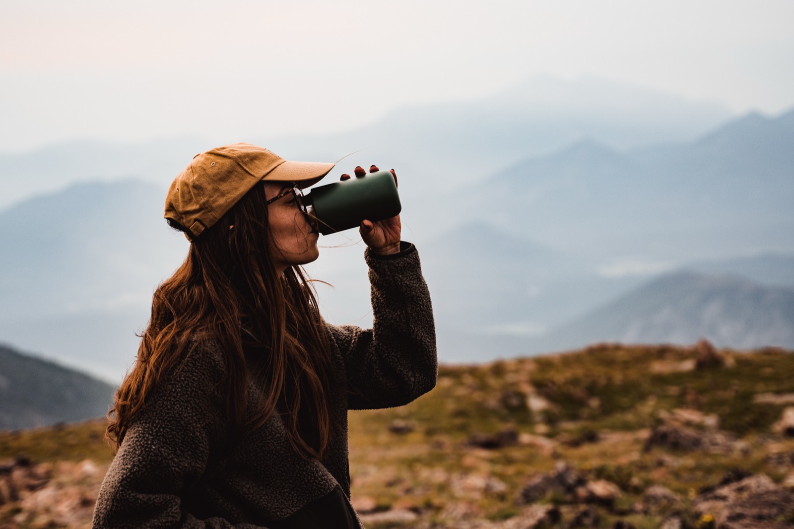 A hiker in an orange beanie drinks from an insulated bottle with hazy mountains behind her.