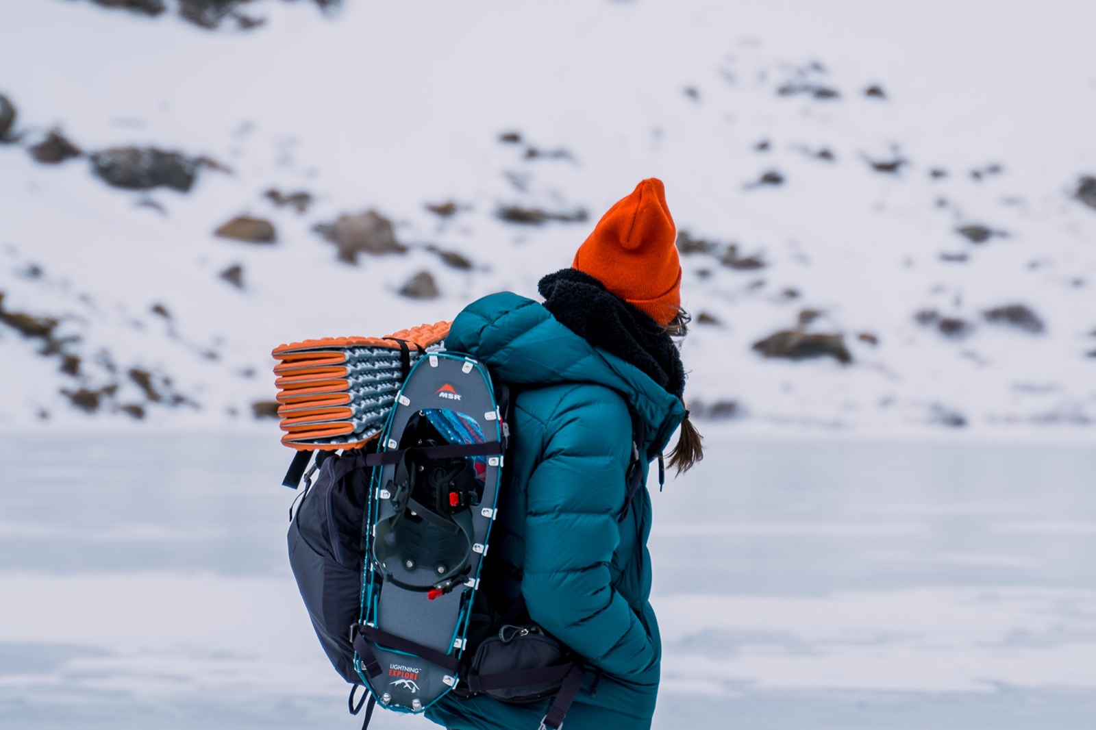 A hiker in an orange beanie carries snowshoes strapped to her pack while walking across a frozen lake.